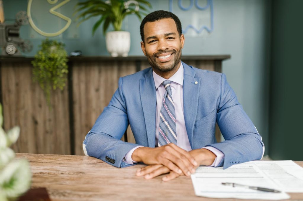 pexels-photo-7821936 Smiling businessman in office attire sitting at a desk with documents.